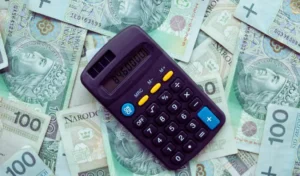 Na zdjęciu widać kalkulator leżący na rozrzuconych banknotach 100 zł. Obraz symbolizuje finanse, liczenie dochodów i zarządzanie budżetem. W kontekście „ile realnie zarabia osoba z orzeczeniem” może odnosić się do wyzwań związanych z dochodami i planowaniem wydatków.